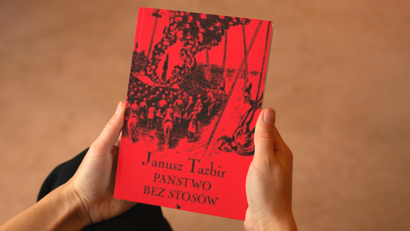 Na brązowym tle widoczne dłonie trzymające książkę Janusza Tazbira "Państwo bez stosów" w jaskrawoczerownej okładce